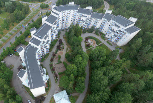 Flygbild av flerbostadshus omgiven av skog med bilväg bredvid.